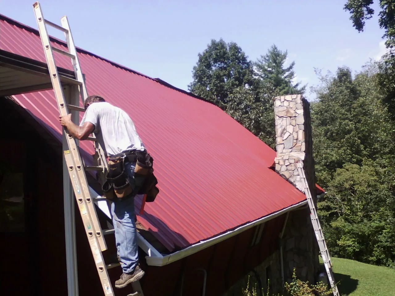 Roofer on a ladder beside a red metal roof and stone chimney — Soffit & Fascia Repair by Roofing Brothers in Sigurd